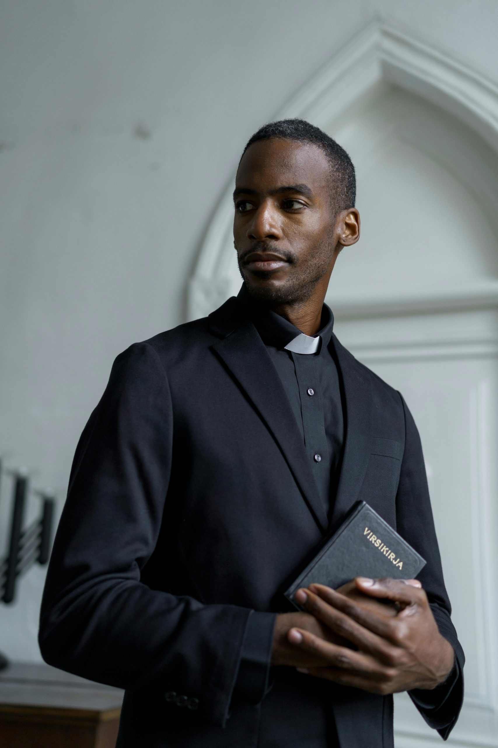 A preacher in a black suit holding a holy book, standing indoors in a church setting.