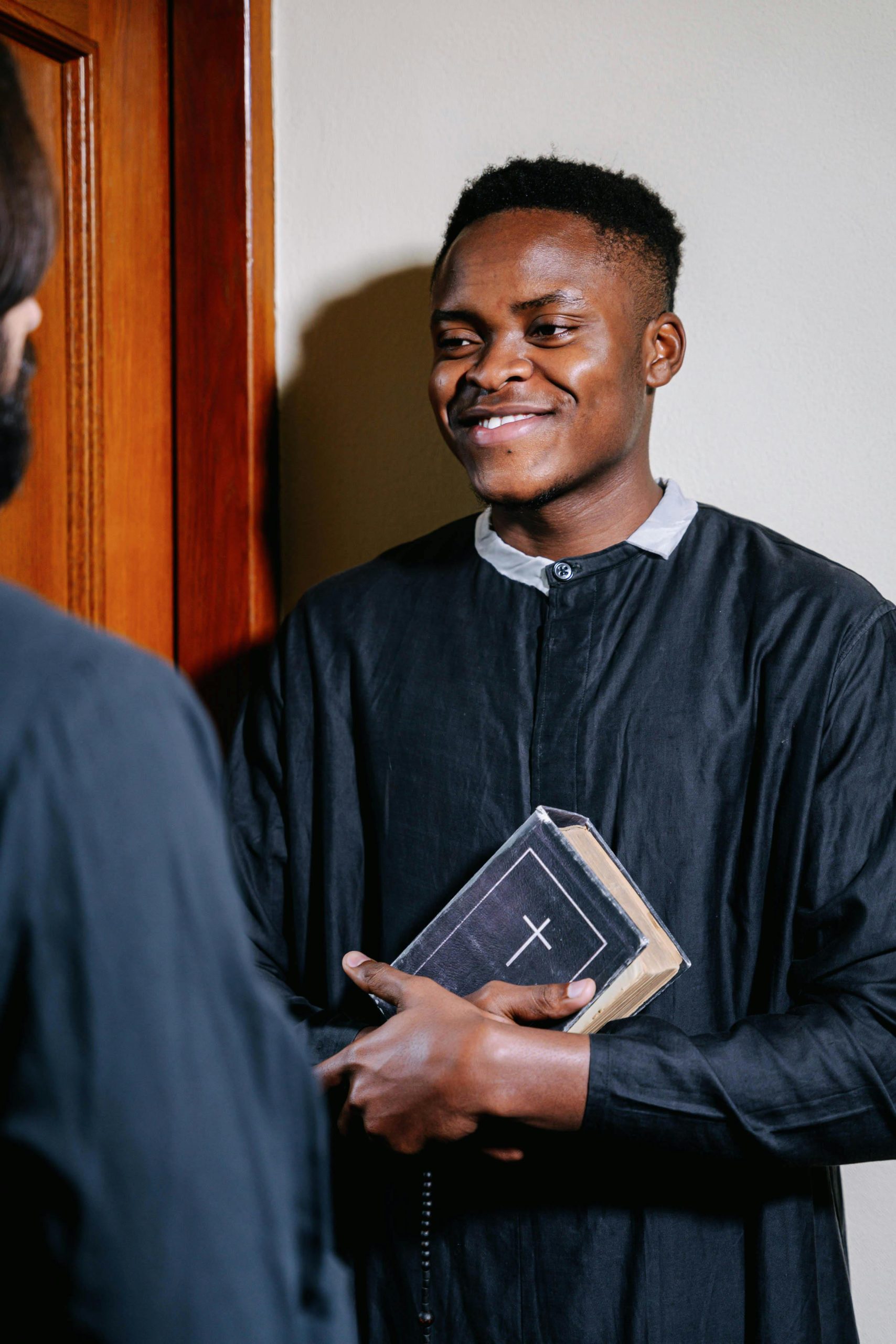Smiling clergyman in a black robe holding a Bible, engaged in a friendly conversation indoors.